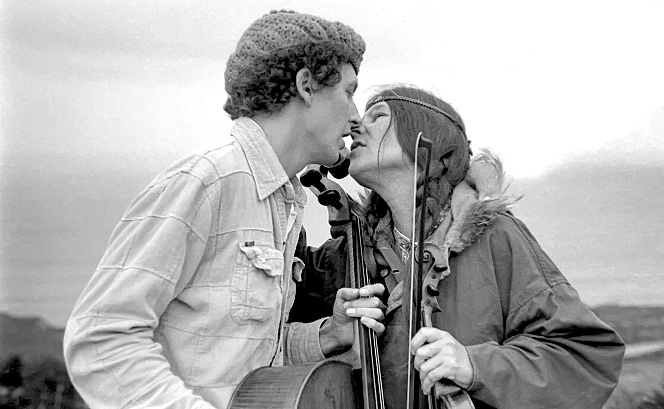 Man and woman with cello and violin almost kiss against a cloudy sky